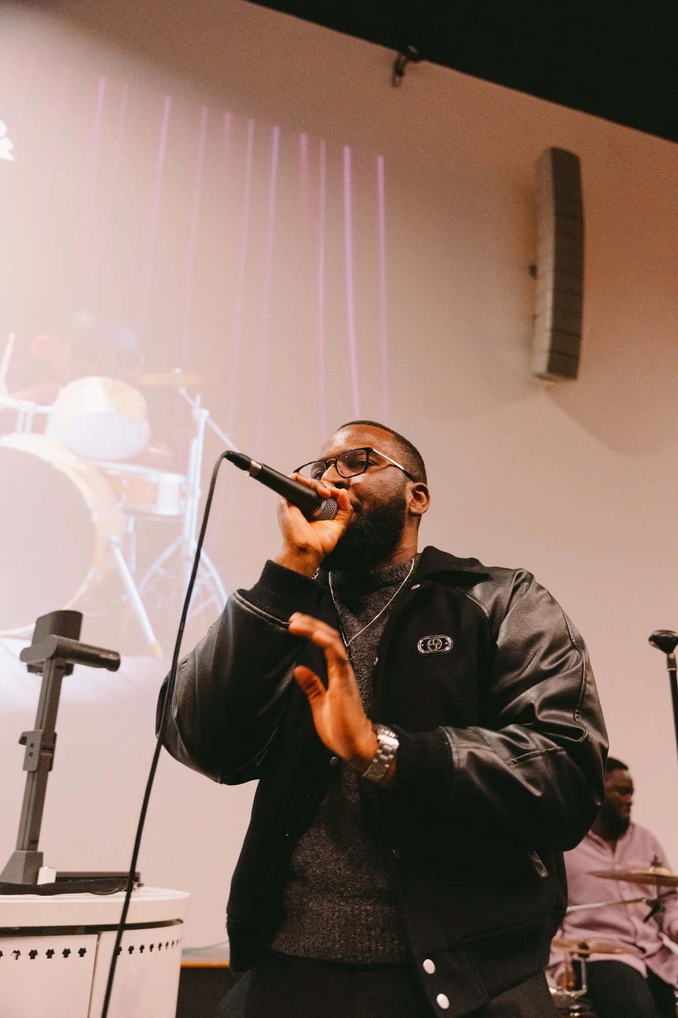 A man with glasses singing into a microphone on stage, with a drummer in the background.