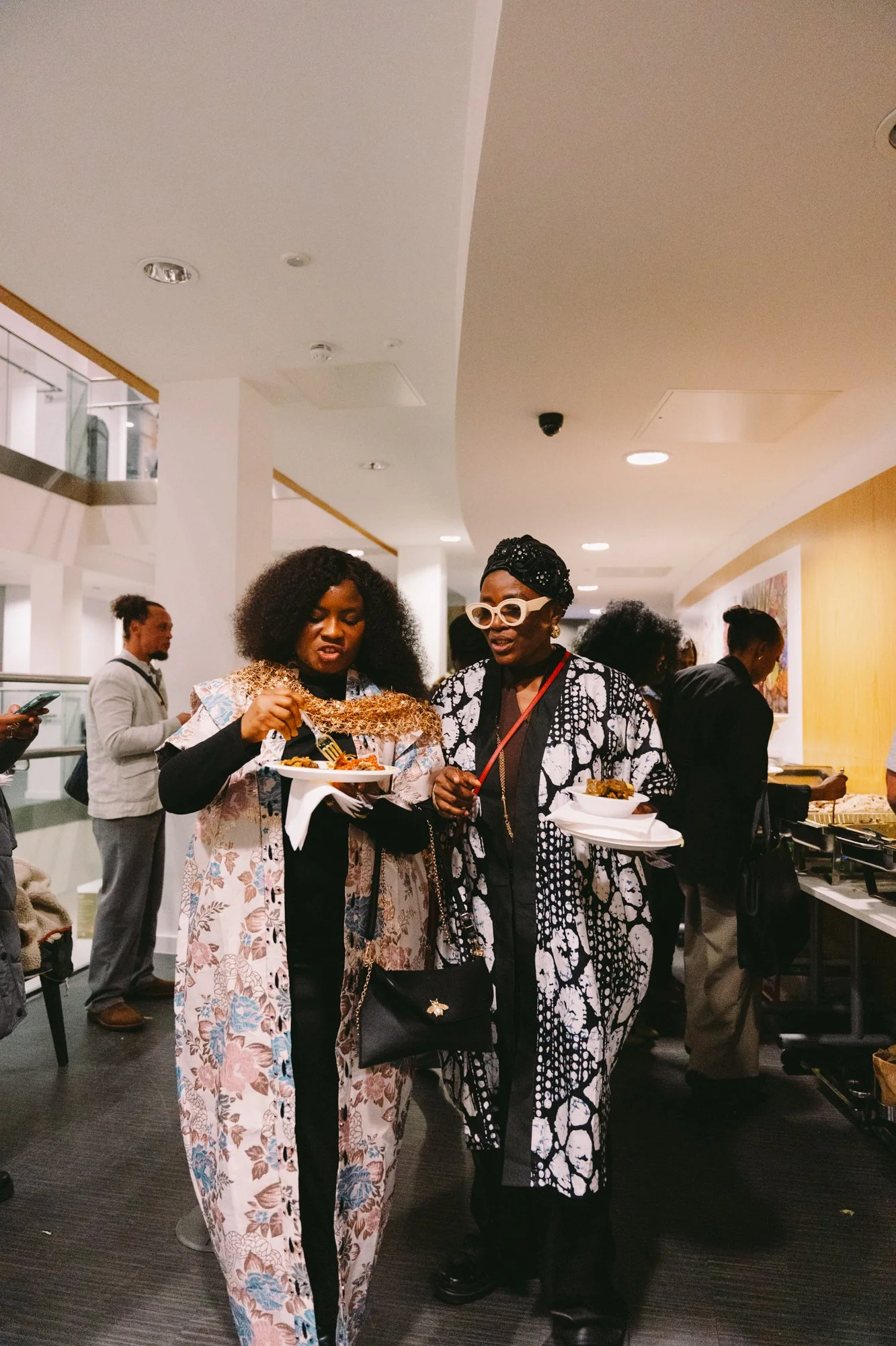 Two women are holding plates of food at a social gathering, standing indoors in a well-lit area with other people in the background.