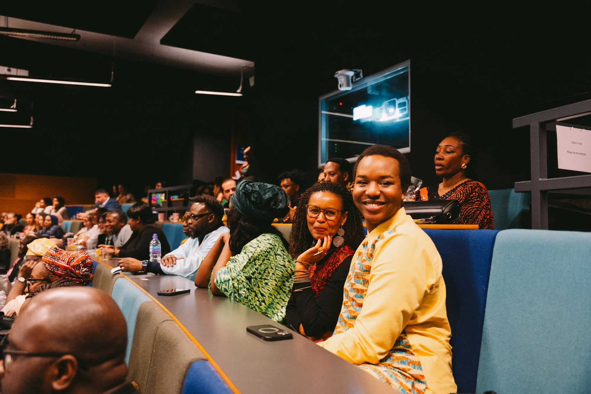 A diverse group of people seated in an auditorium, engaged and smiling, with a focus on three individuals in the front row.