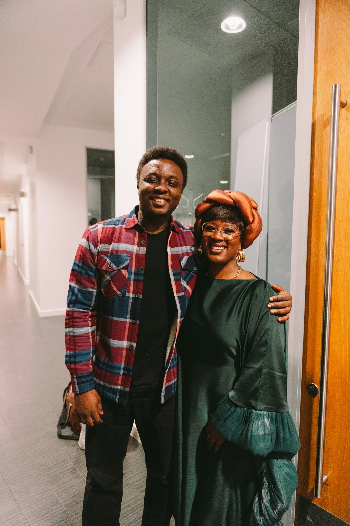 A man and a woman smiling, standing close together indoors. The man wears a red and blue plaid shirt over a black T-shirt, and the woman wears a dark green dress with puffed sleeves and a large orange headwrap. She also wears glasses and gold jewelry