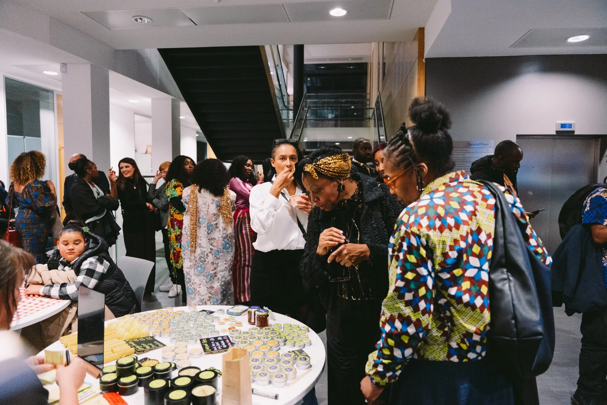 A diverse group of people gathered around a table with various small jars and containers in a modern indoor setting. Some individuals are looking at products, others are browsing, and a few are chatting. The background features a staircase and elevat