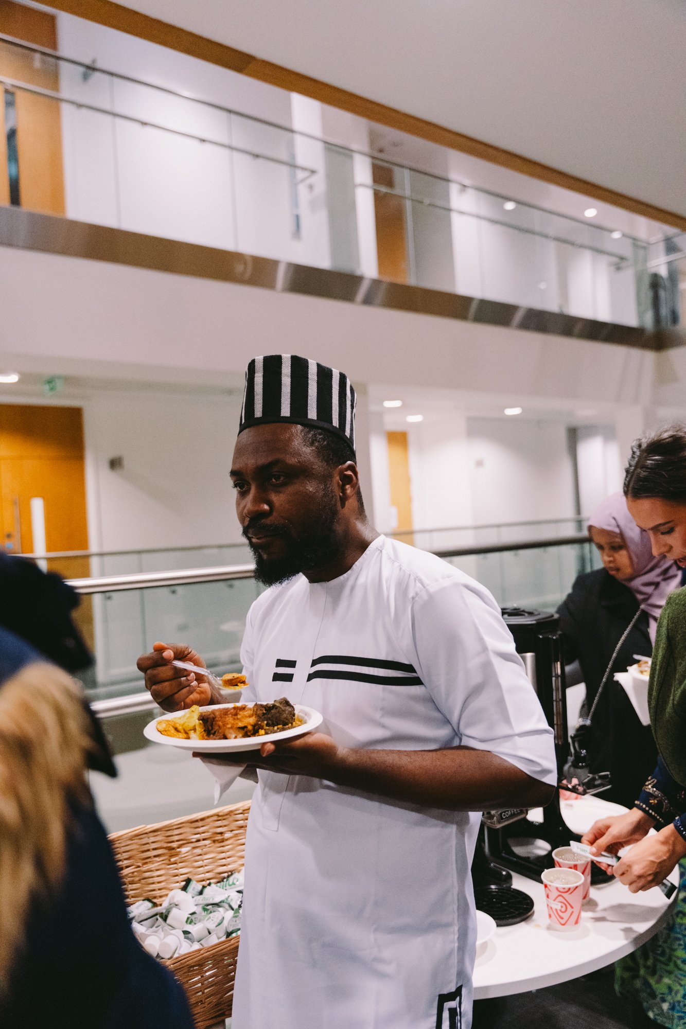 A chef in a white uniform and a black-and-white striped chef hat holding a plate of food at a buffet, with people in the background serving themselves.