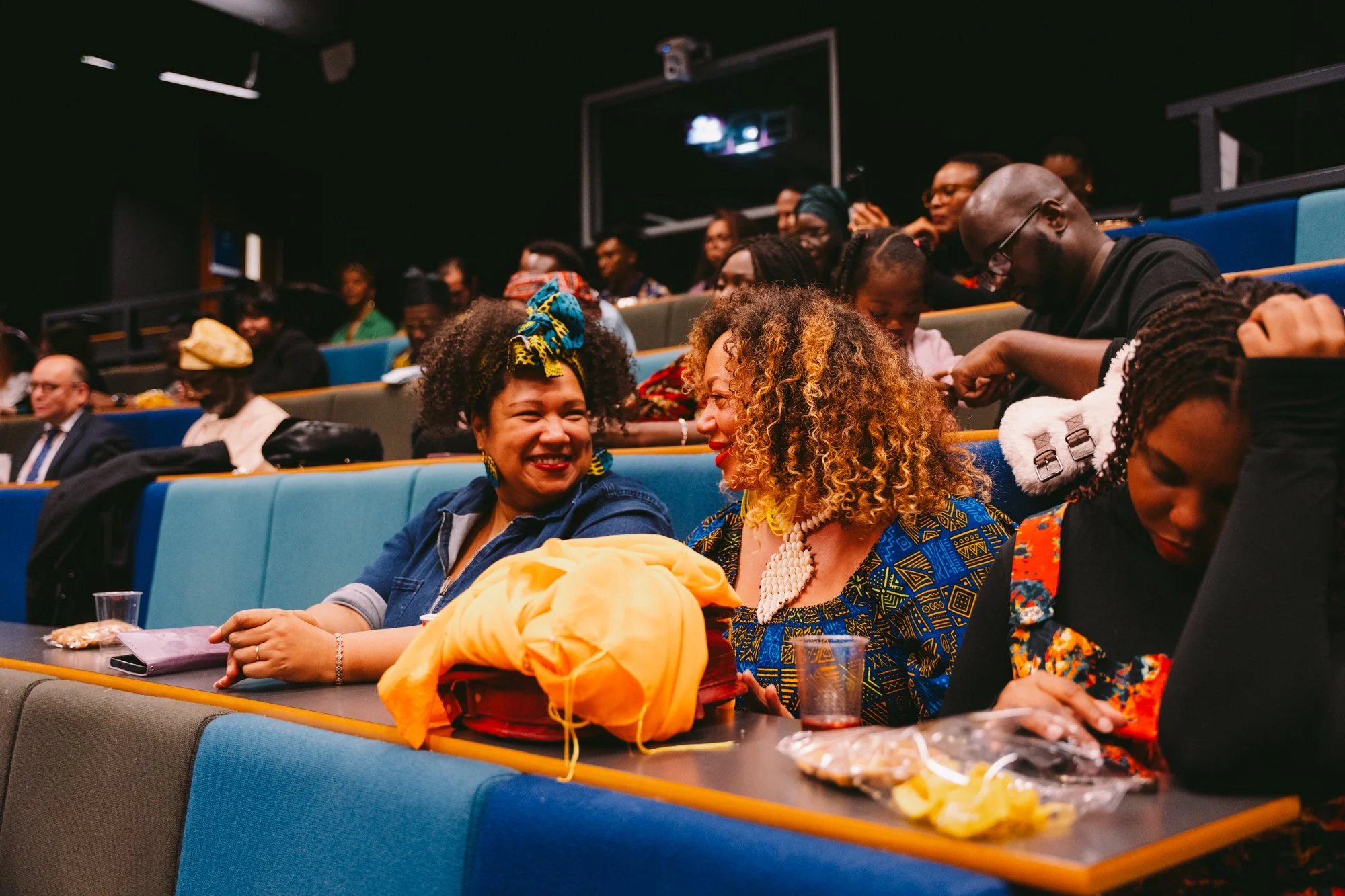 People sitting in a theater or auditorium, engaging in conversation and smiling, with some holding snacks or drinks.