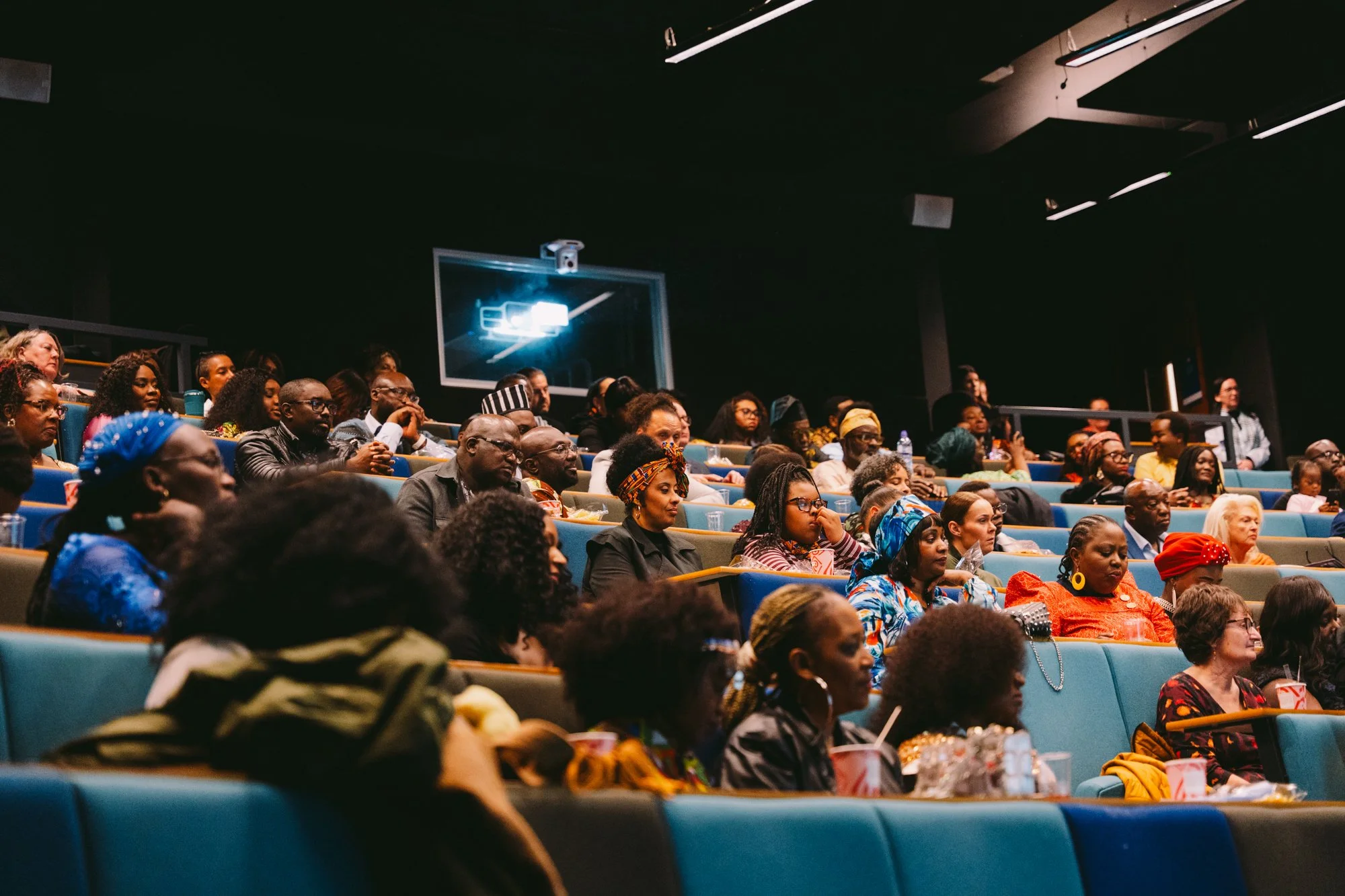 Audience seated in theater watching a presentation, with various colorful clothing and headscarves.