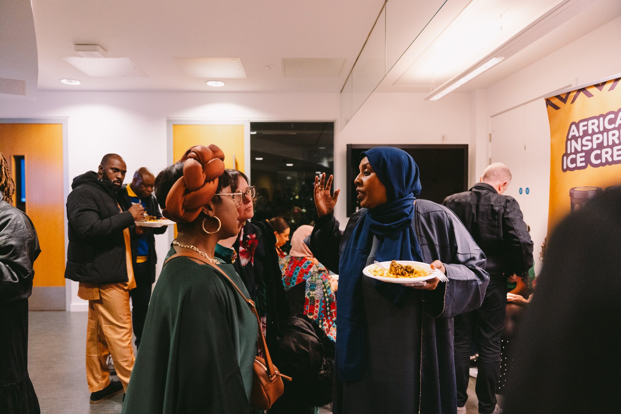 Group of people at a social gathering indoors, engaging in conversation with a woman in a blue hijab gesturing while holding a plate of food.