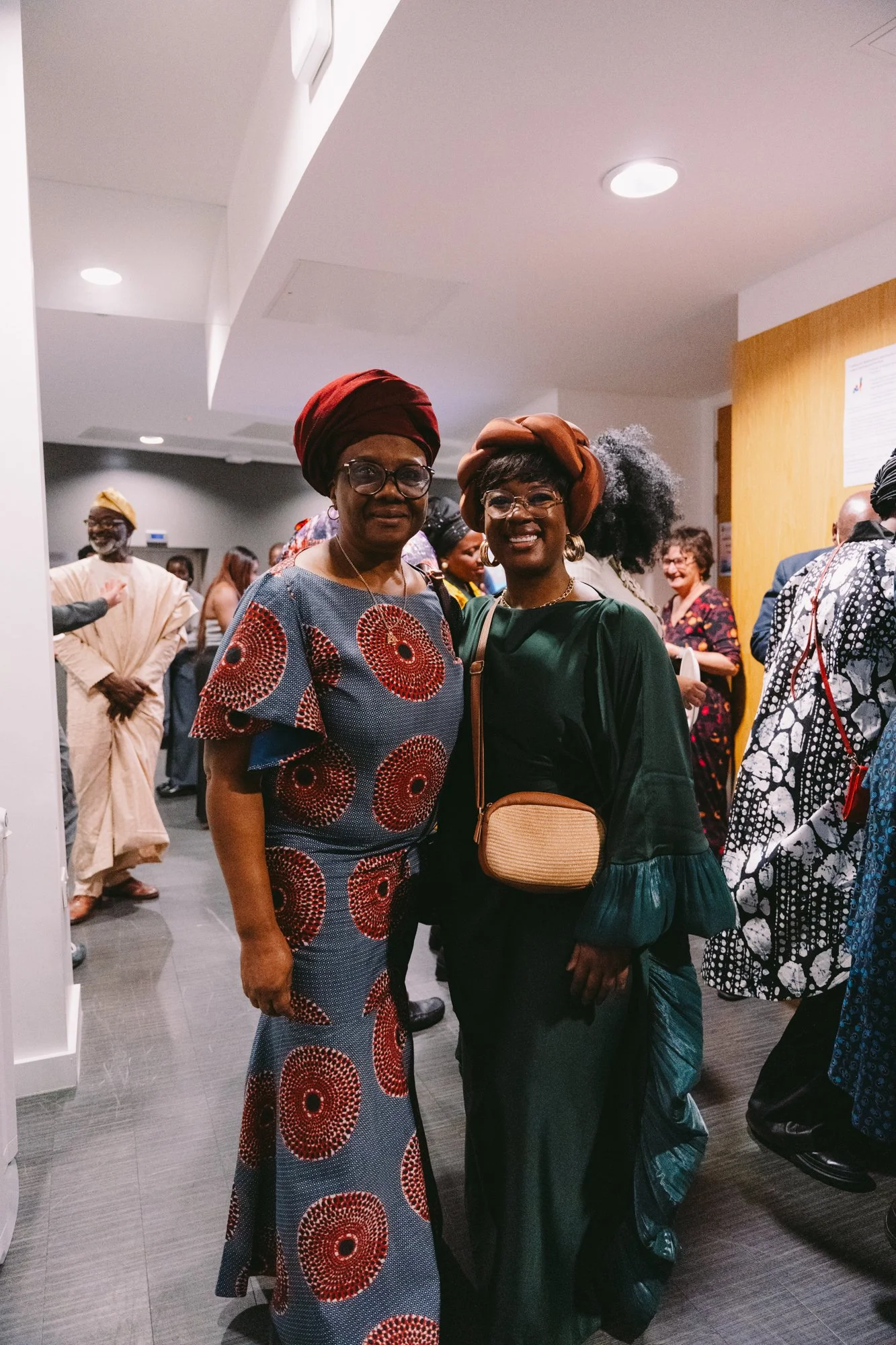 Two women at an indoor event, dressed in traditional African clothing and accessories, standing and smiling in front of a group of people in the background.