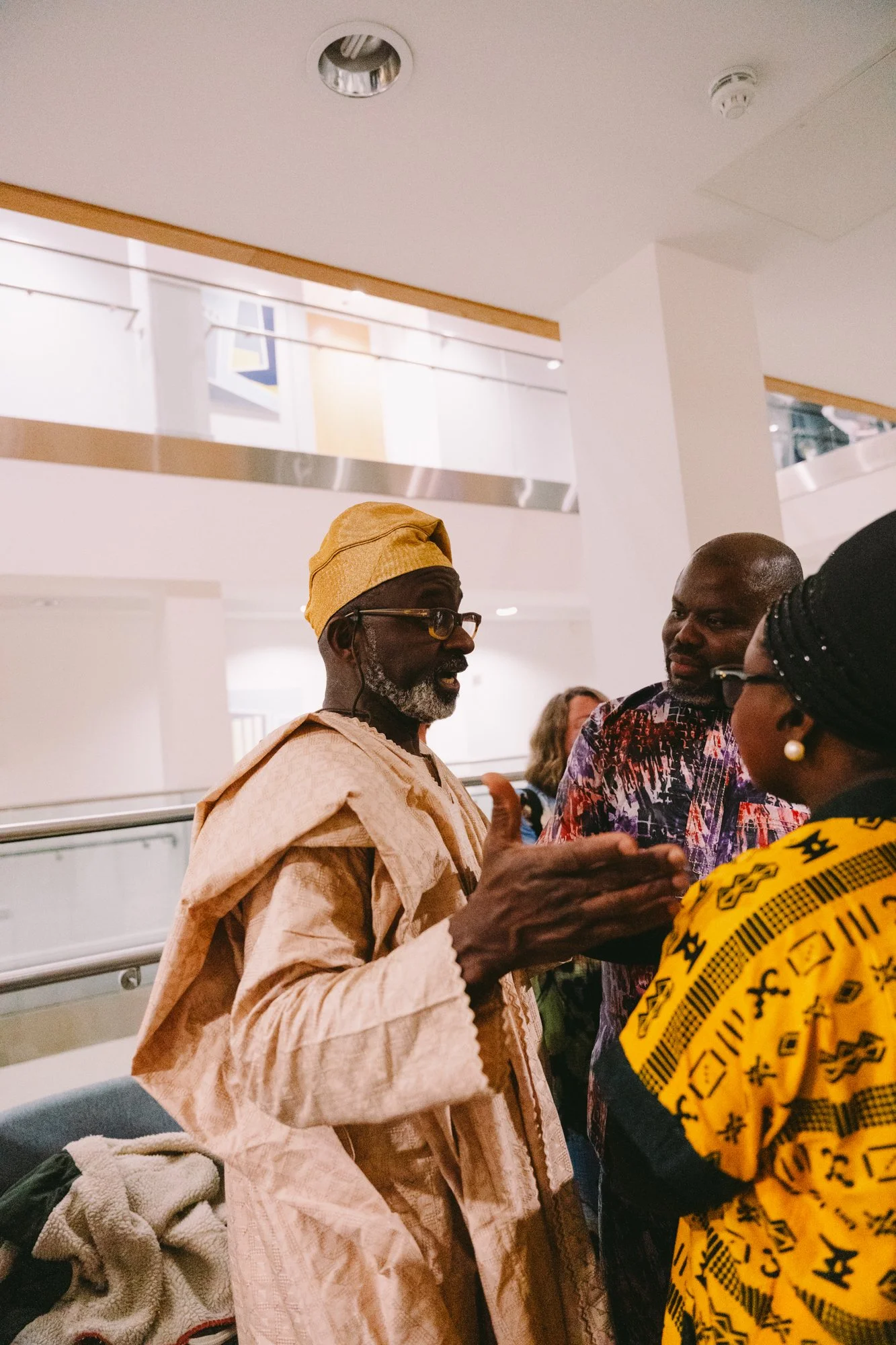 Three people engaged in conversation indoors, with one man wearing traditional African clothing and a headwrap, speaking to a woman with glasses and a headscarf, and another man observing. Background includes a white wall and upper balcony area.
