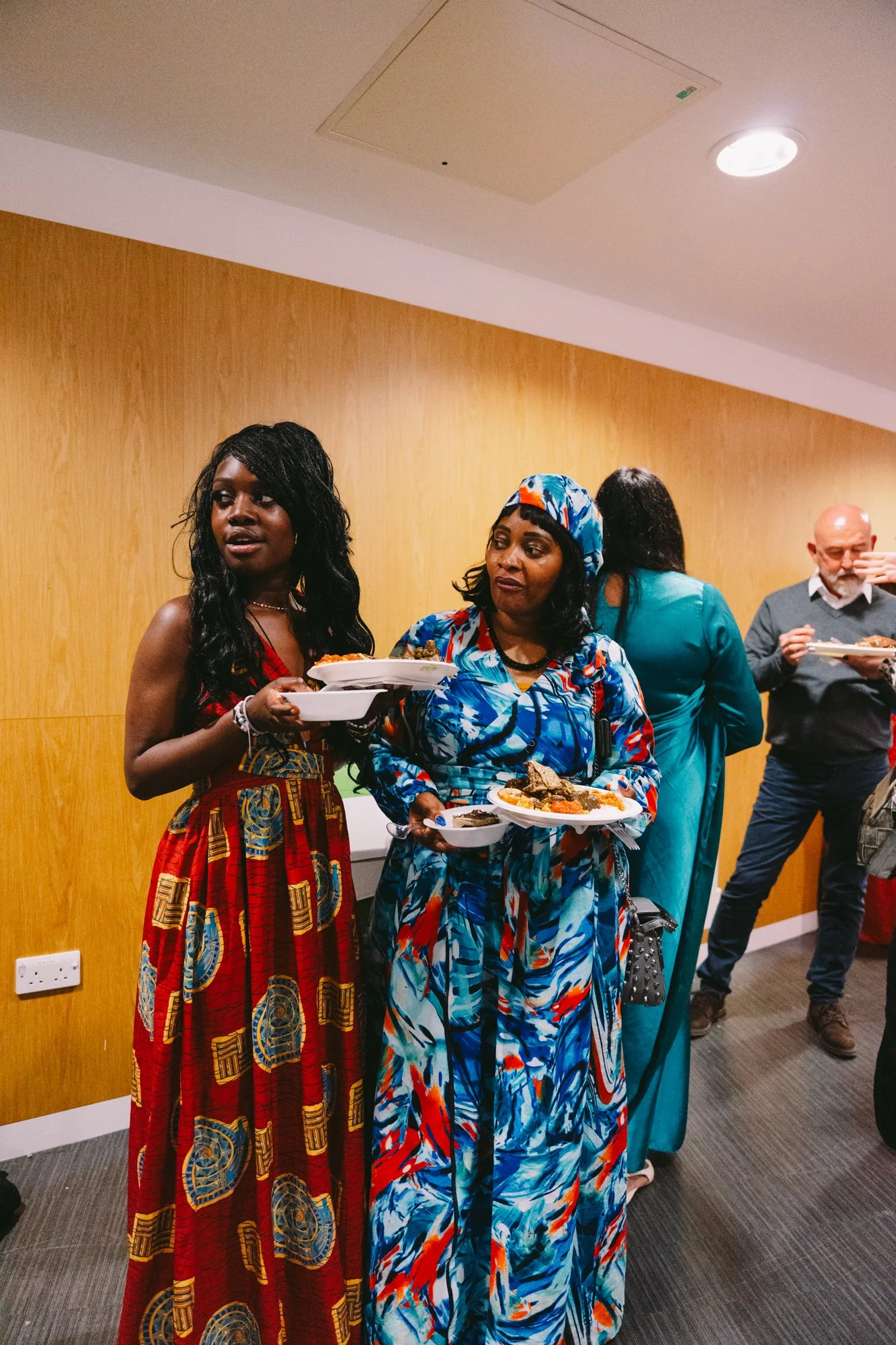 Three women and two men in a room, holding plates of food at a social gathering. The women are dressed in colorful dresses, and the men are in casual attire, standing against a wood-paneled wall with a ceiling vent and lights.