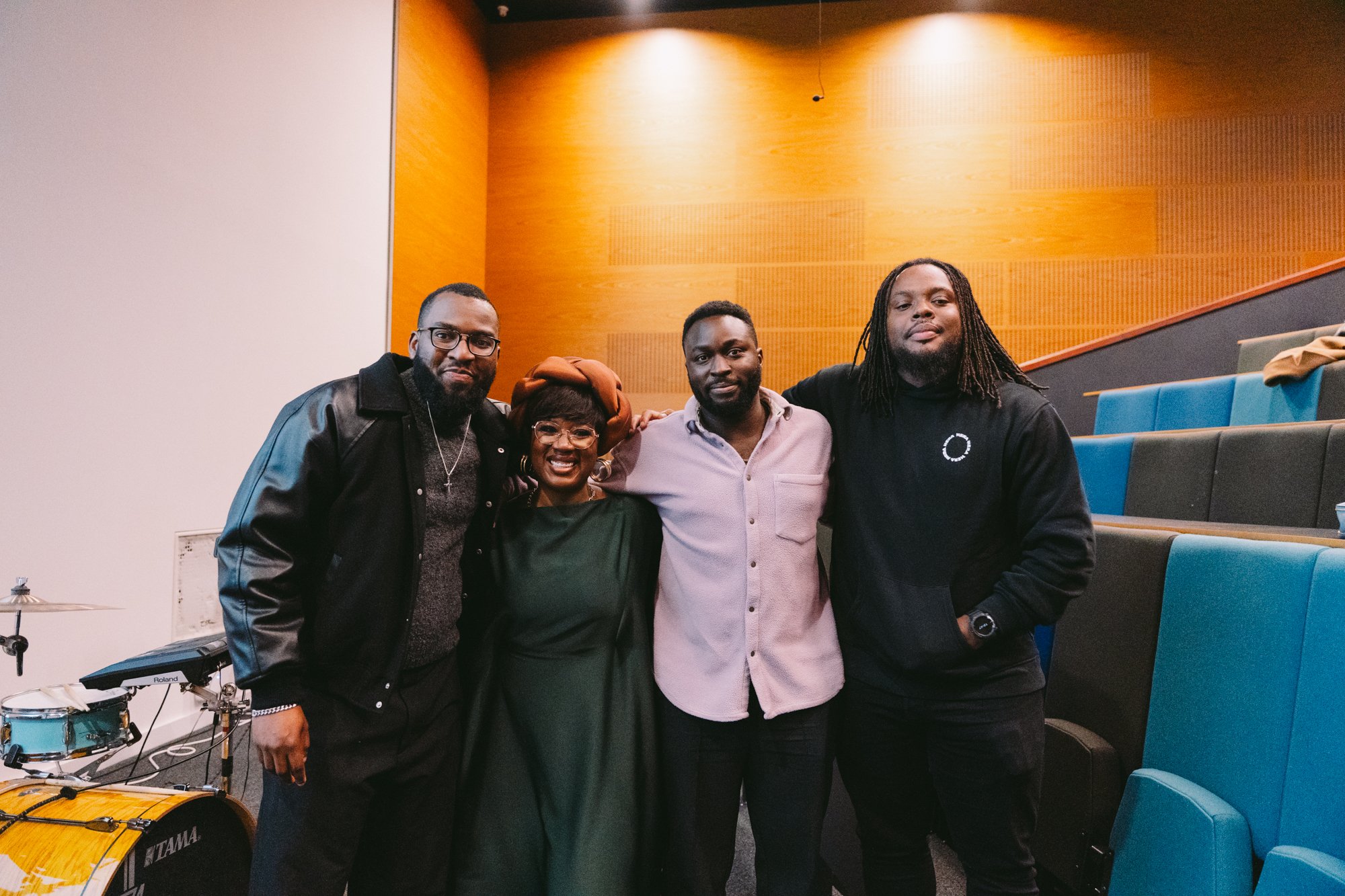 Four people standing together inside a room with blue and gray tiered seating. They are smiling, with one person's arm around another, and appear to be posing for a group photo. To the left, there is a drum set and electronic keyboard.