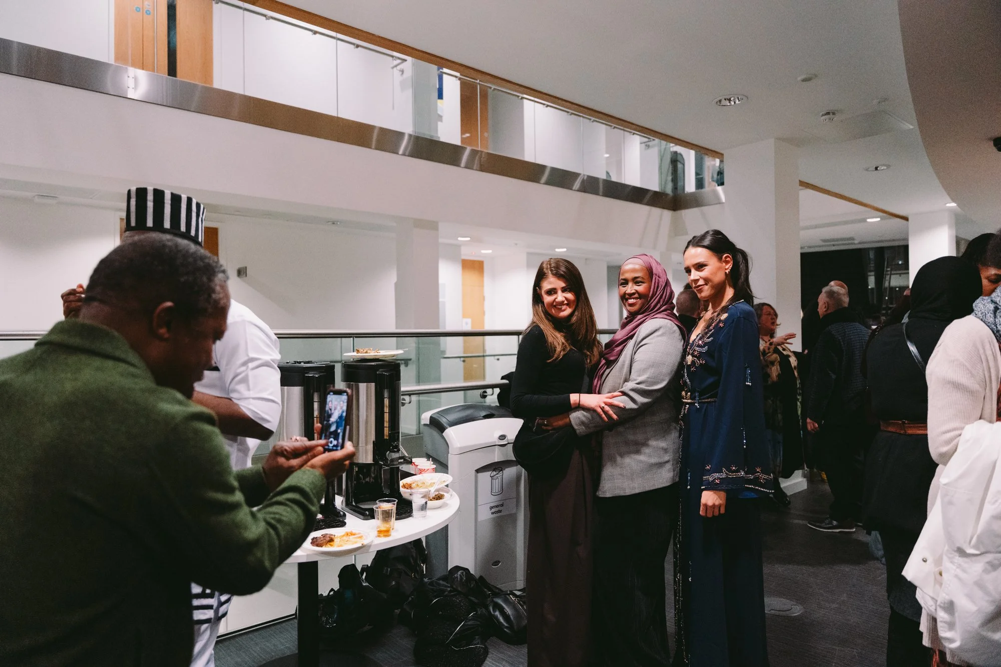 Three women stand close together, smiling, at a social event. One woman is wearing a black top, another a gray blazer with a hijab, and the third a dark blue dress with embroidery. In the foreground, a person in a green jacket is taking a picture wit