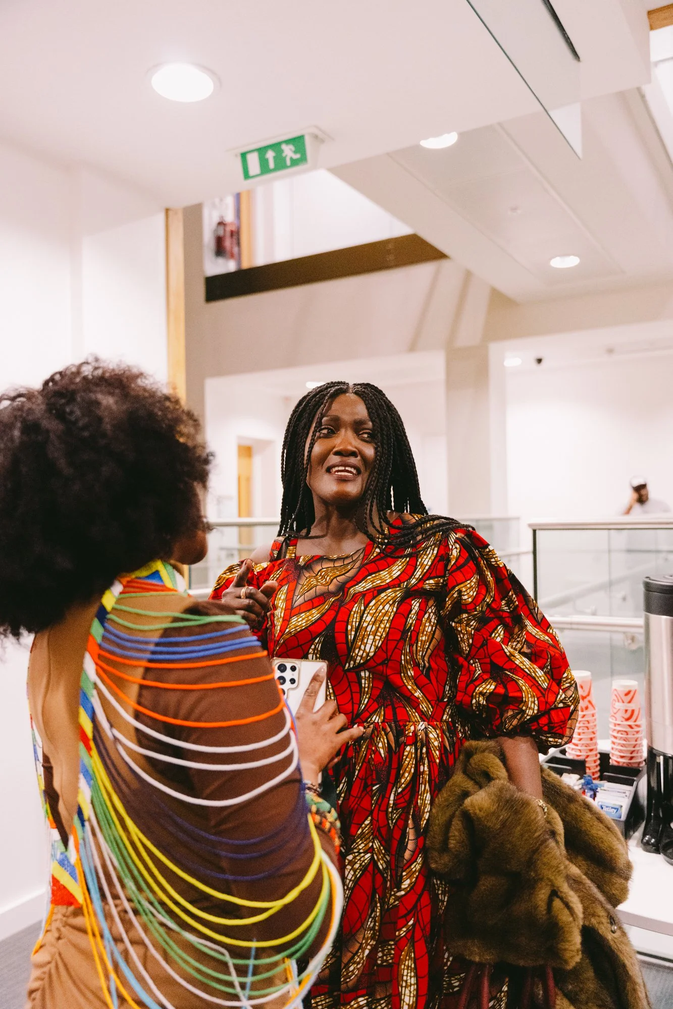 Two women talking in an indoor setting, one wearing a colorful dress with a fur coat over her arm, the other wearing a rainbow-striped top, with a beverage station in the background.