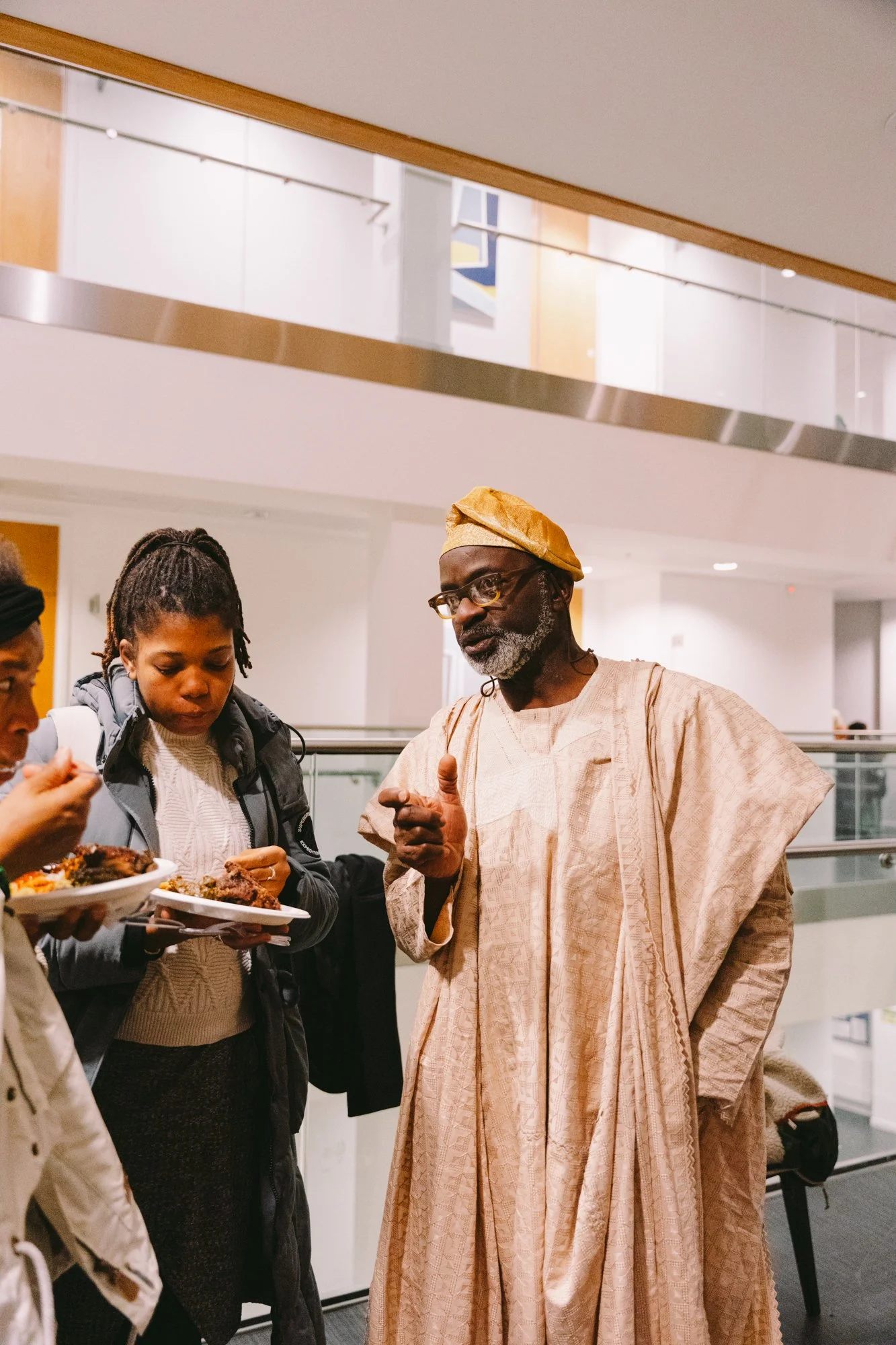 An older man wearing traditional African attire and a yellow cap is talking to two young women, one of whom is holding a plate of food, in an indoor space.