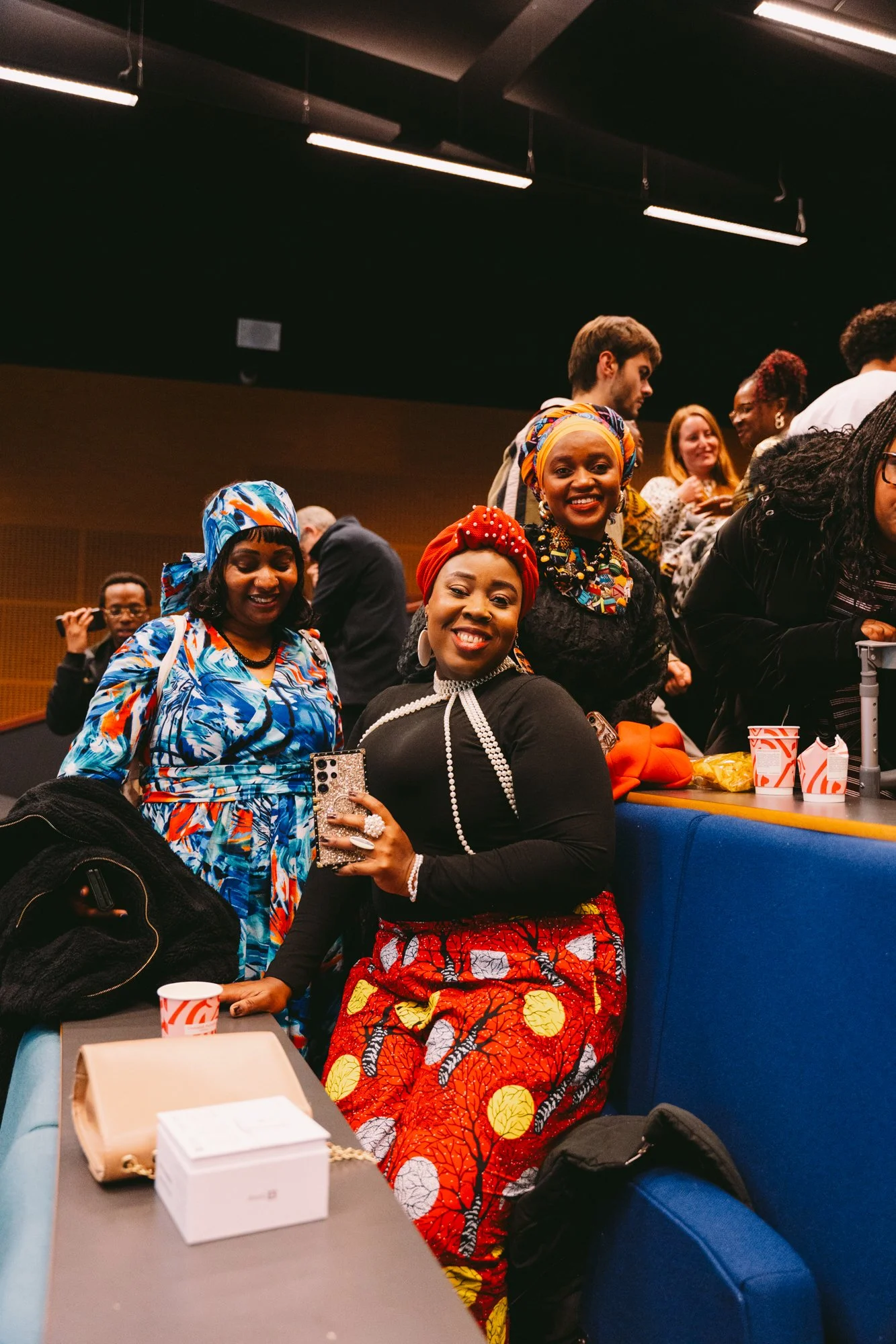 Group of women and men at a social event, with three women in colorful headwraps and skirts in the foreground, smiling and enjoying themselves.