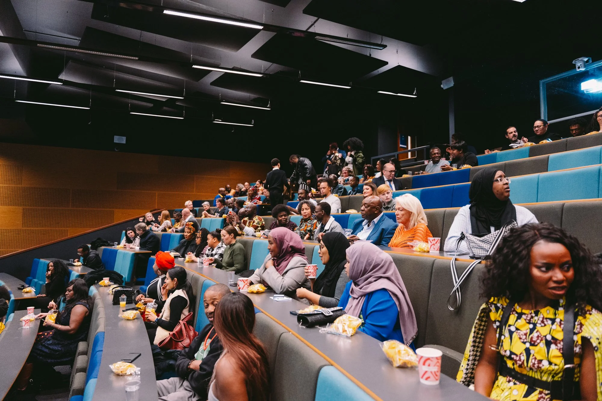 A diverse group of people seated in an auditorium, some engaged in conversation, eating snacks, or using their phones, with a few standing, in a modern indoor setting with dark ceiling and wooden wall panels.