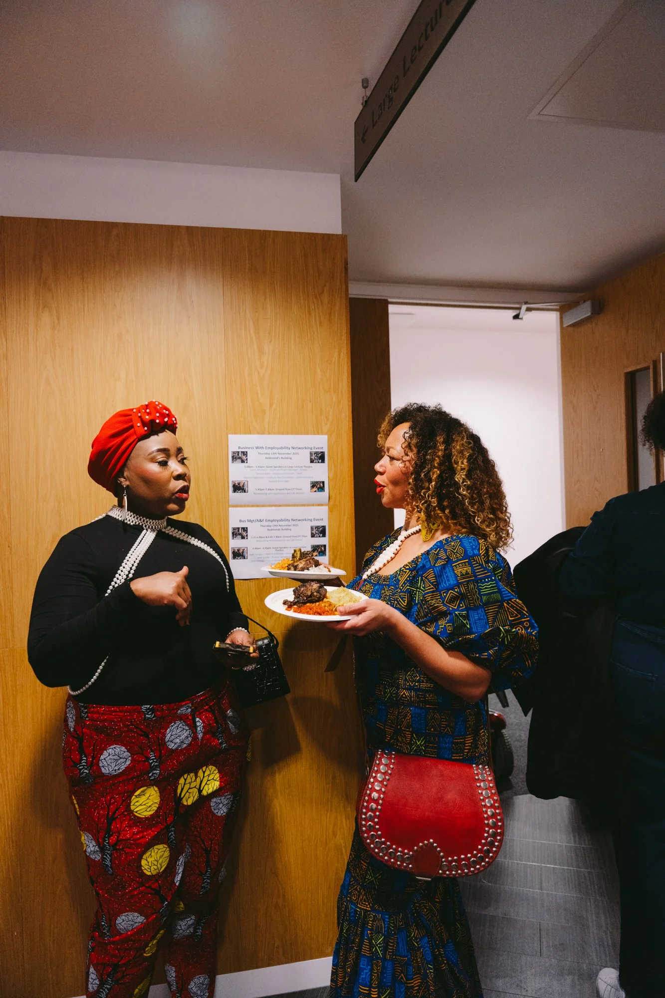 Two women engaged in conversation at an event, holding plates of food, standing indoors near a wooden wall with event posters.