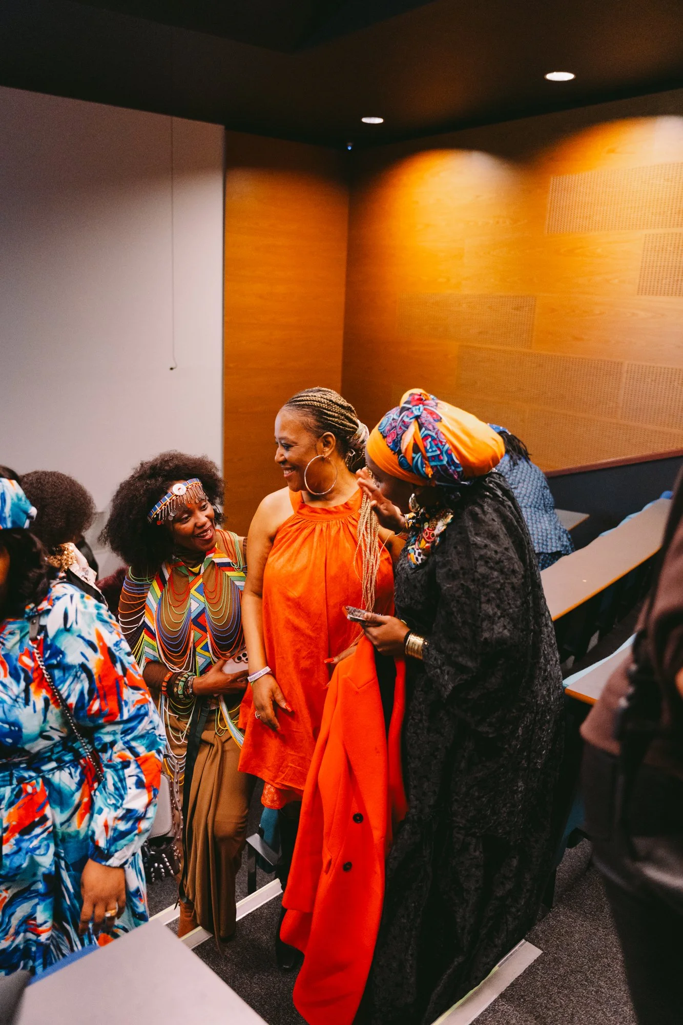 A group of women dressed in colorful and traditional African attire, smiling and engaging in conversation in a well-lit room with wood-paneled walls.