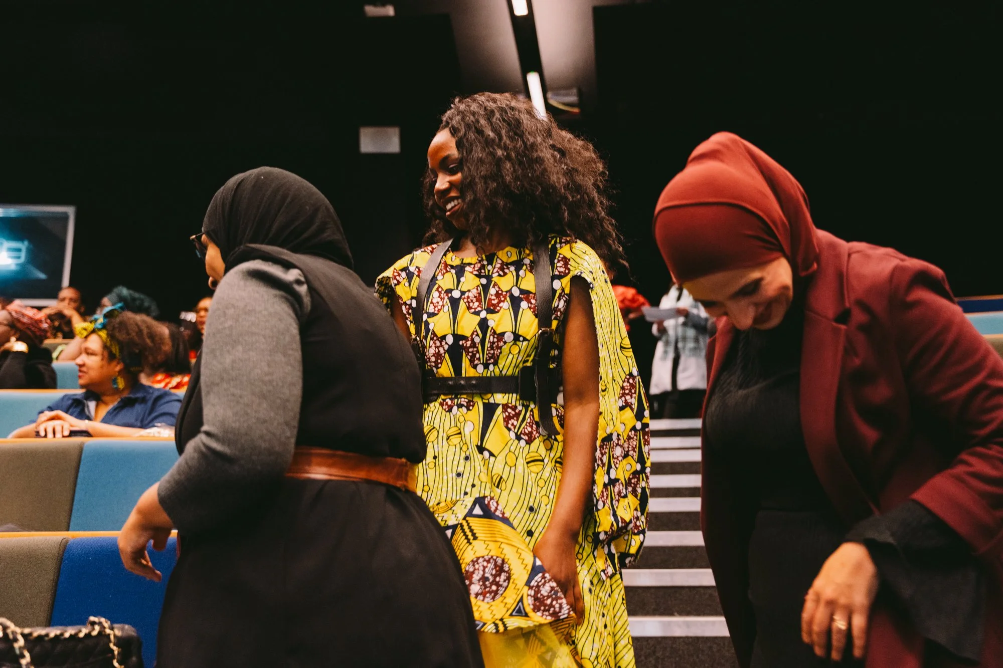 Three women smiling and talking in an auditorium. One woman wears a black and gray jacket with a belt, another wears a brightly colored patterned dress, and the third wears a maroon headscarf and matching blazer.