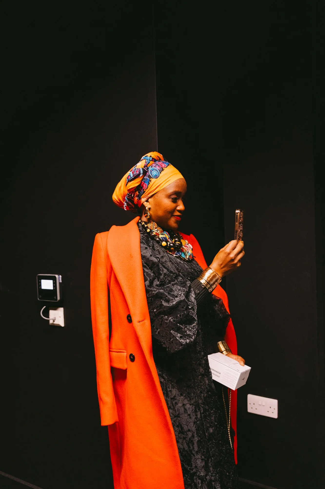 Woman wearing a colorful headwrap and an orange coat, standing against a dark wall, looking at her phone.