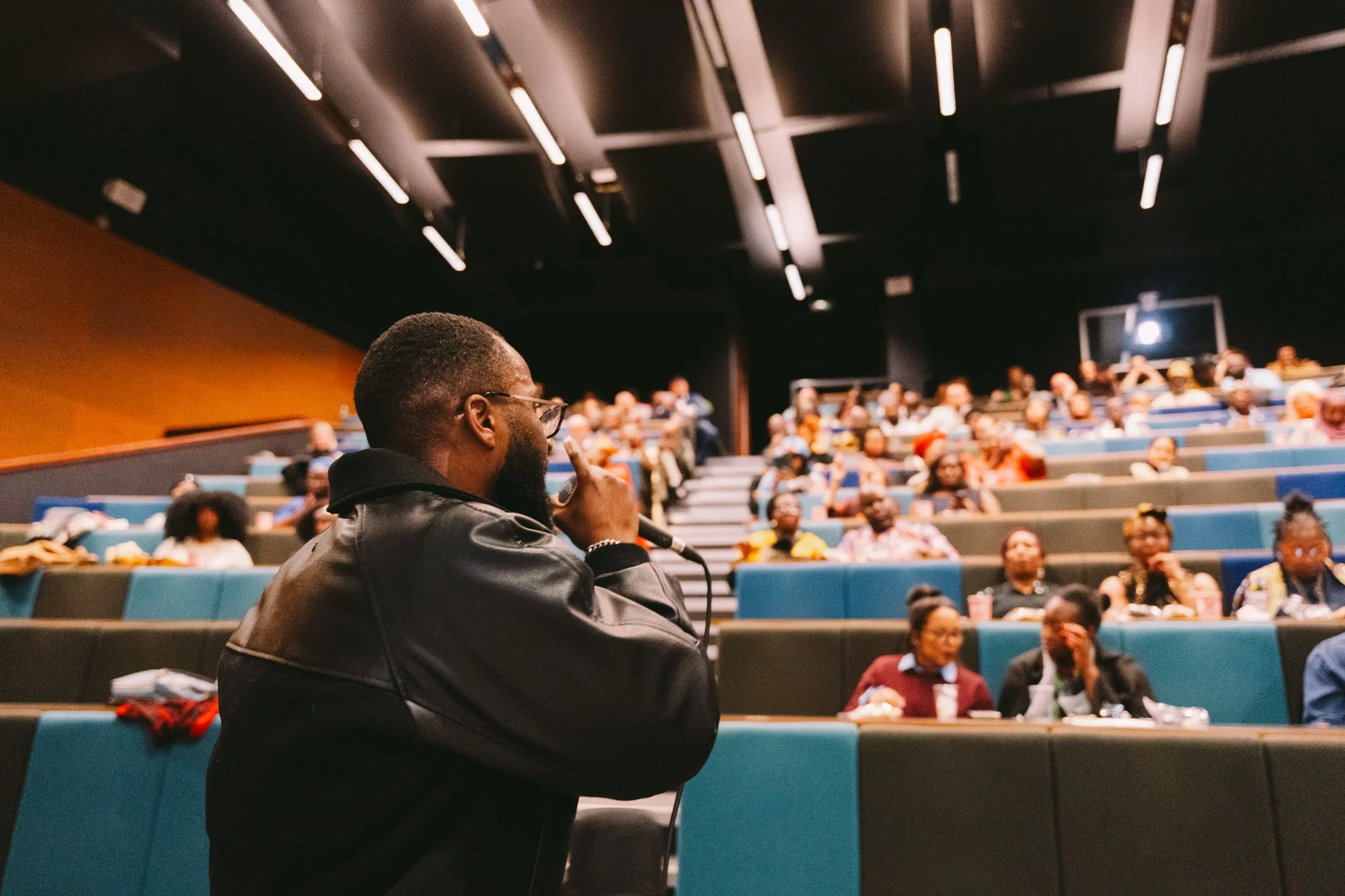 A man with glasses holding a microphone near his mouth is speaking to an audience in an auditorium with tiered seating and a dark ceiling.
