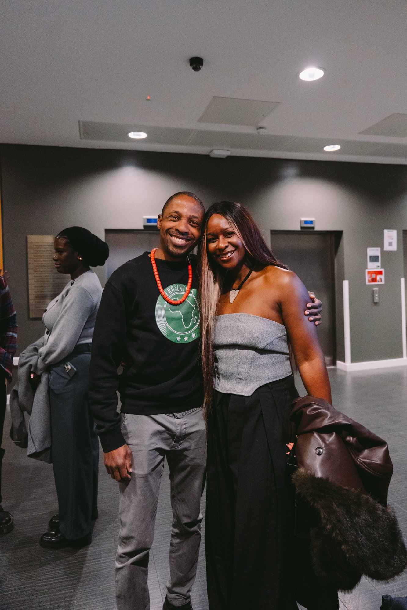 Two people smiling and posing for a photo in an indoor setting, with others in line in the background.