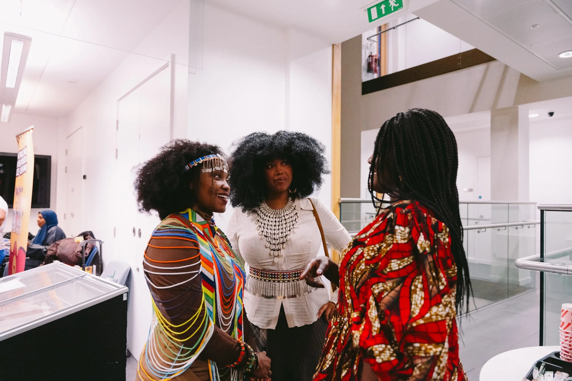 Three women with black curly hair, dressed in colorful traditional African clothing, are engaged in conversation indoors, with a modern interior setting.