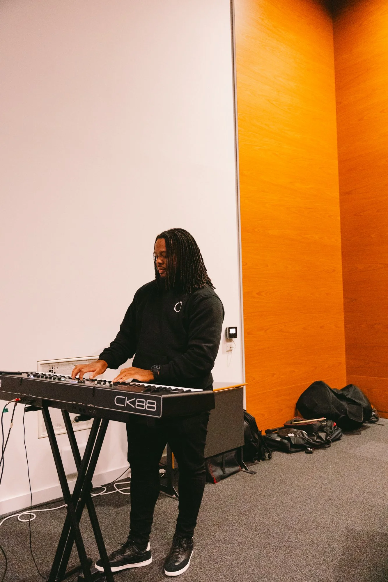 A man with long dreadlocks playing a keyboard in a room with beige and wooden walls.