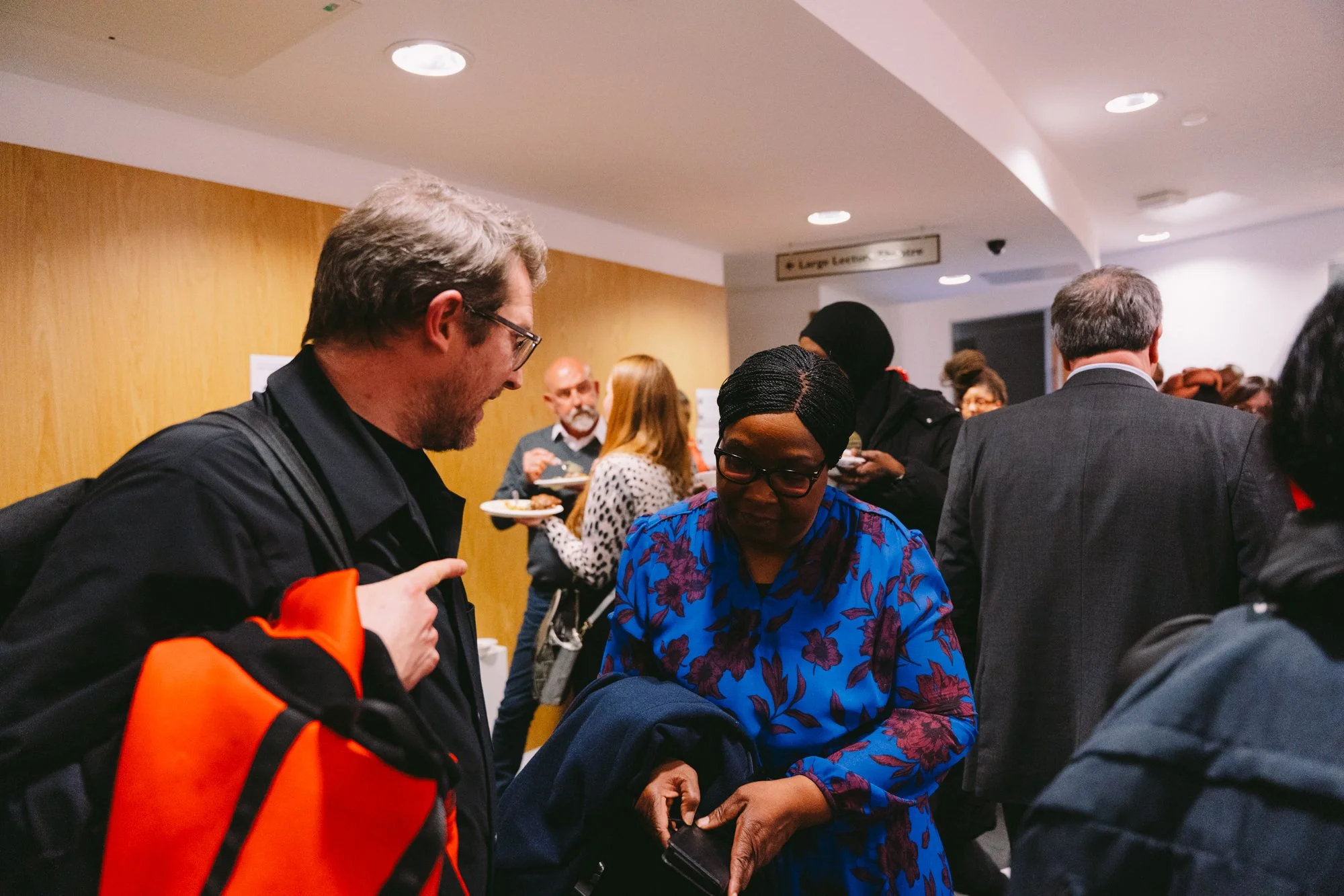 People in a crowded indoor space, some holding plates of food, engaging in conversations. The background includes a wooden wall and a sign pointing to a nearby location.