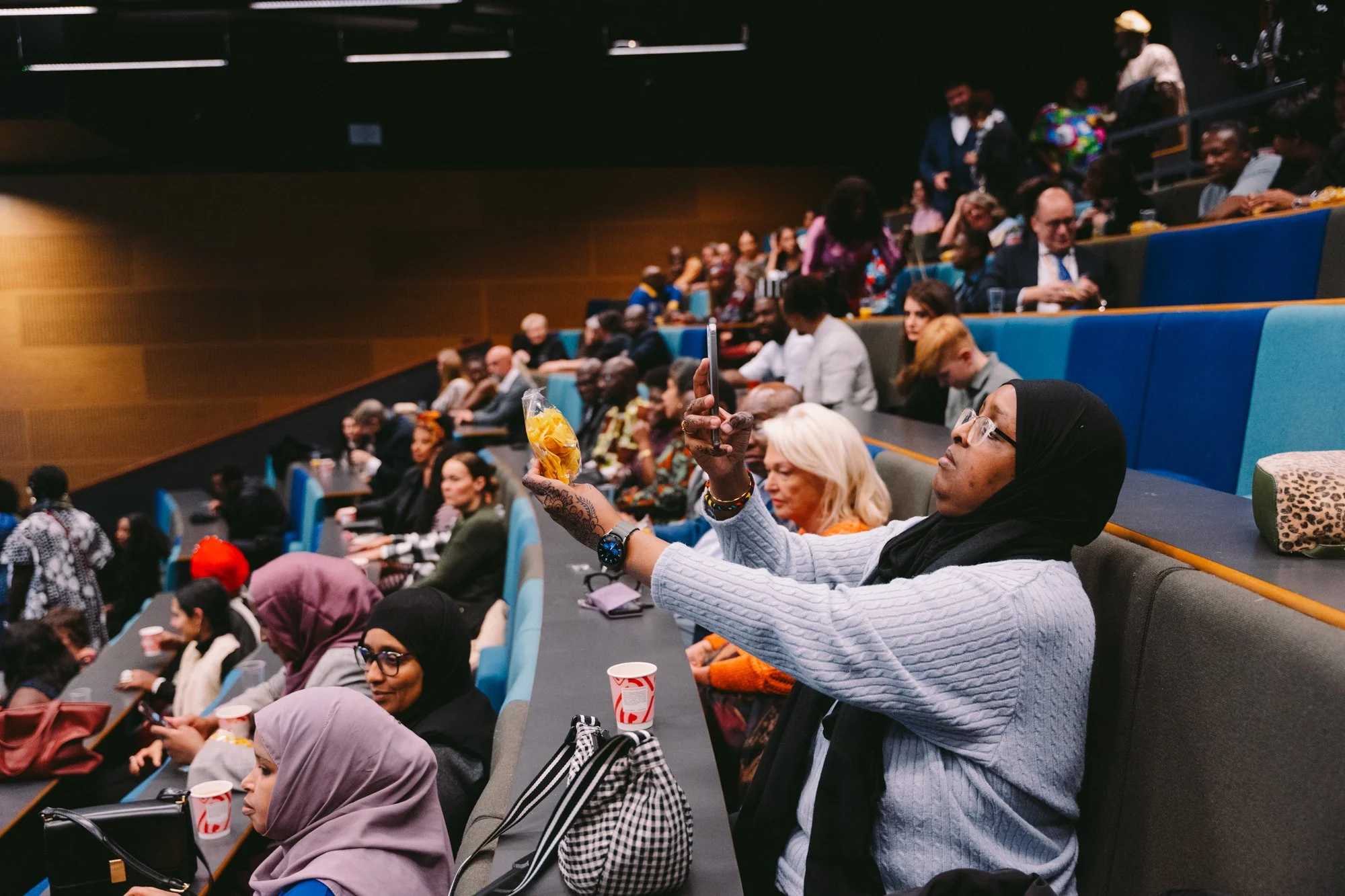 Audience seated in an auditorium, some taking photos, wearing diverse attire including hijabs, suits, and casual clothing.