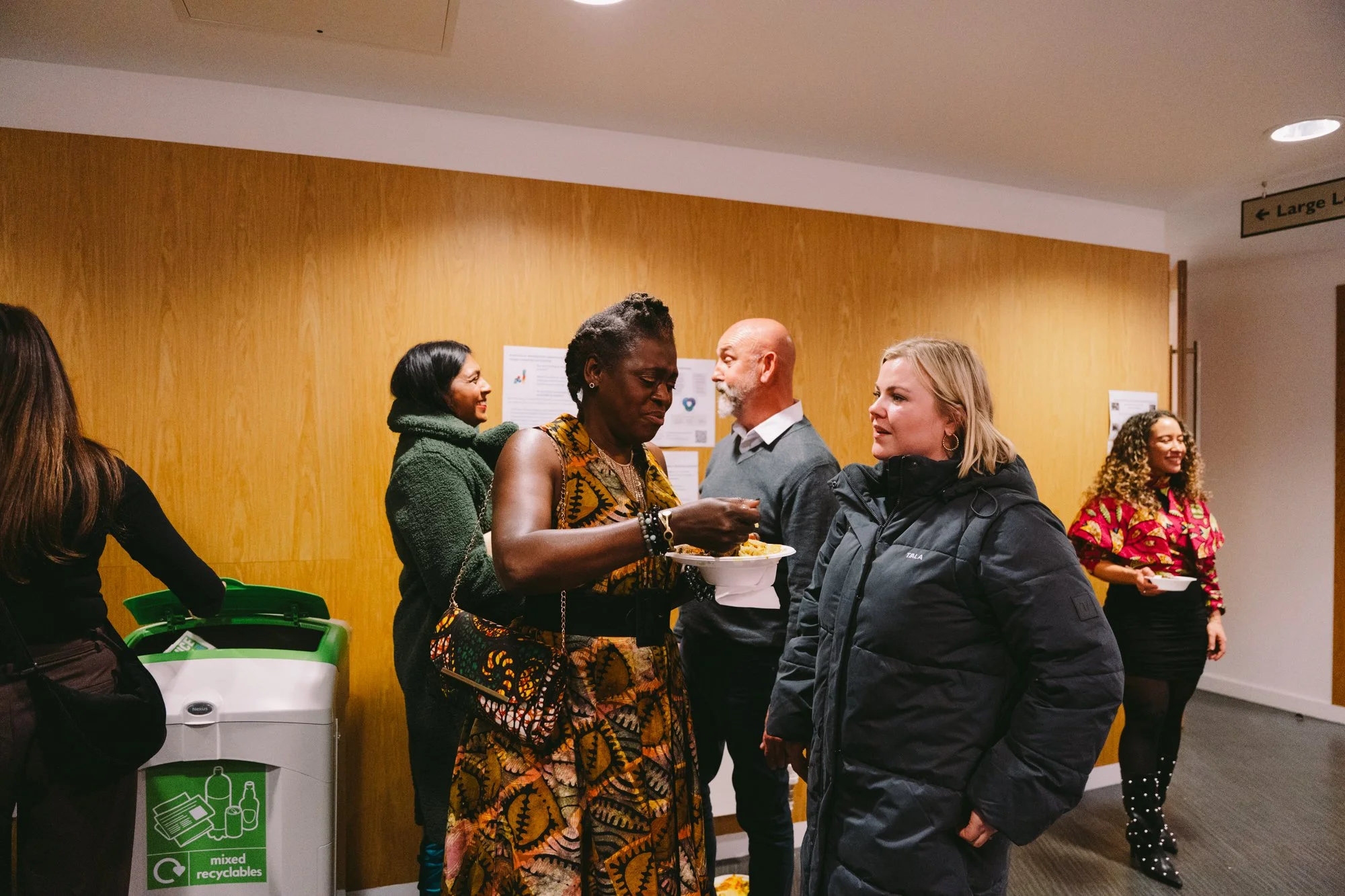 A group of diverse people socializing at an indoor event, engaged in conversation, with some holding food plates, against a wood-paneled wall.