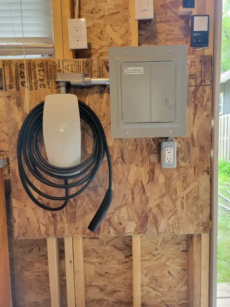 Electric vehicle charging station with a coiled charging cable, electrical panel, and outlet mounted on a plywood wall in a garage or workshop.