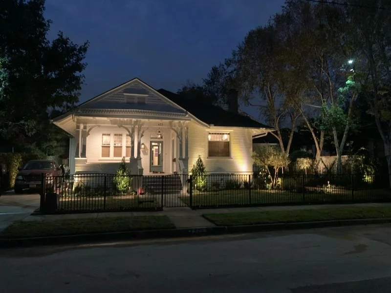 A well-lit white house with a front porch, surrounded by a black metal fence, trees, and a lawn at night.