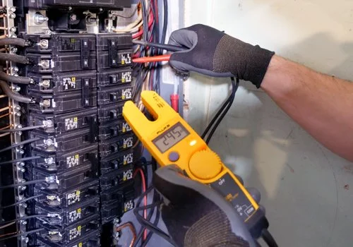 Person using a digital multimeter to test electrical wiring inside an electrical panel.