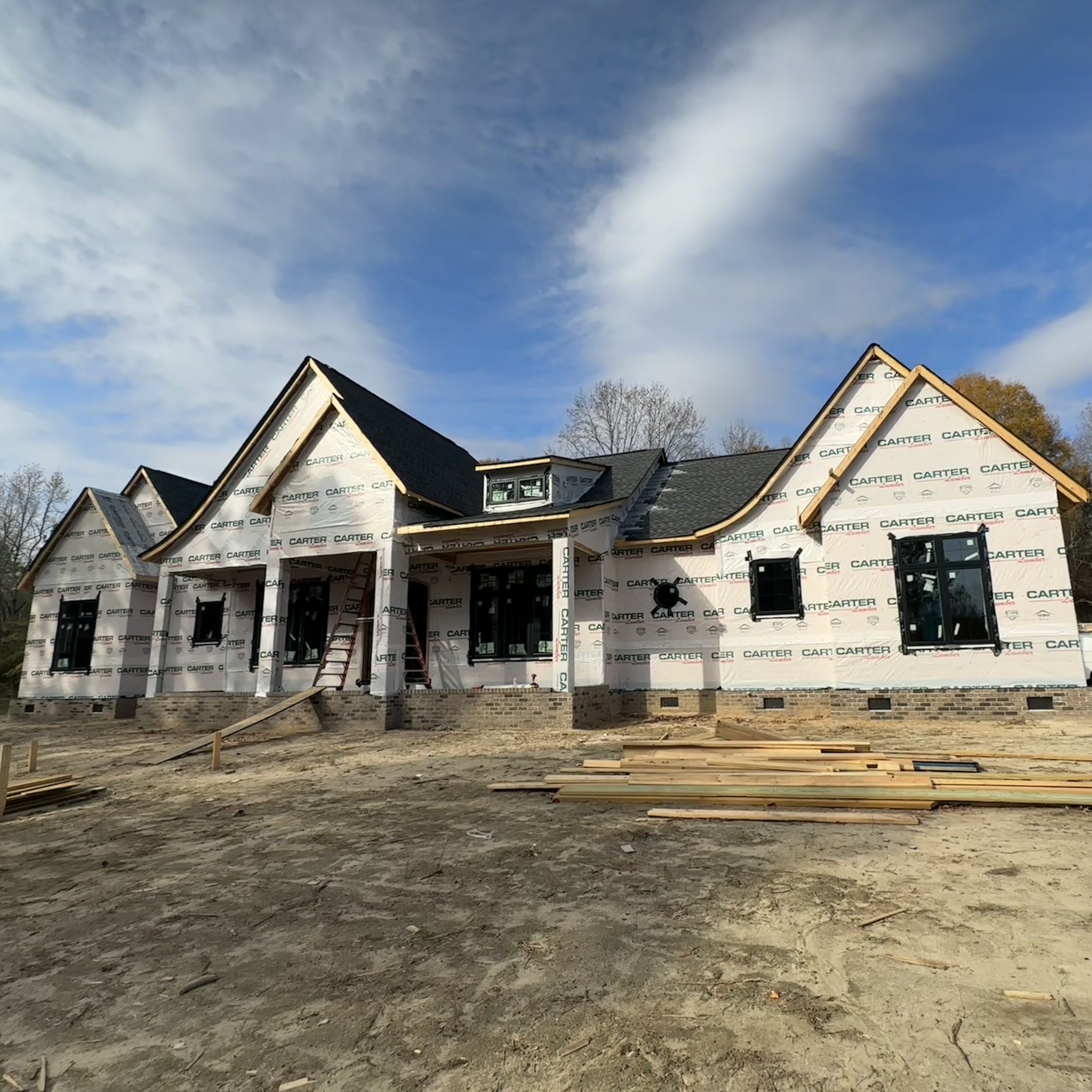 Under-construction house wrapped in building materials with visible windows and a peaked roof under a partly cloudy sky.