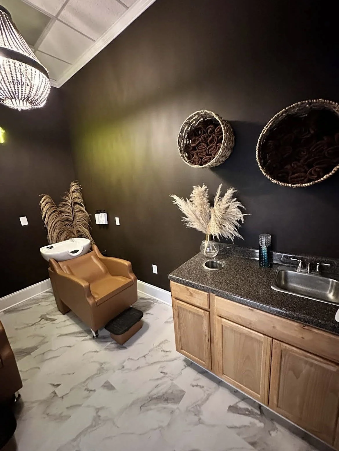 A salon or spa setting with a brown leather chair, a sink, and a granite countertop with a vase of pampas grass. The dark wall has woven baskets with rolled towels and a decorative lamp hanging from the ceiling. The floor is marble with gray veining.