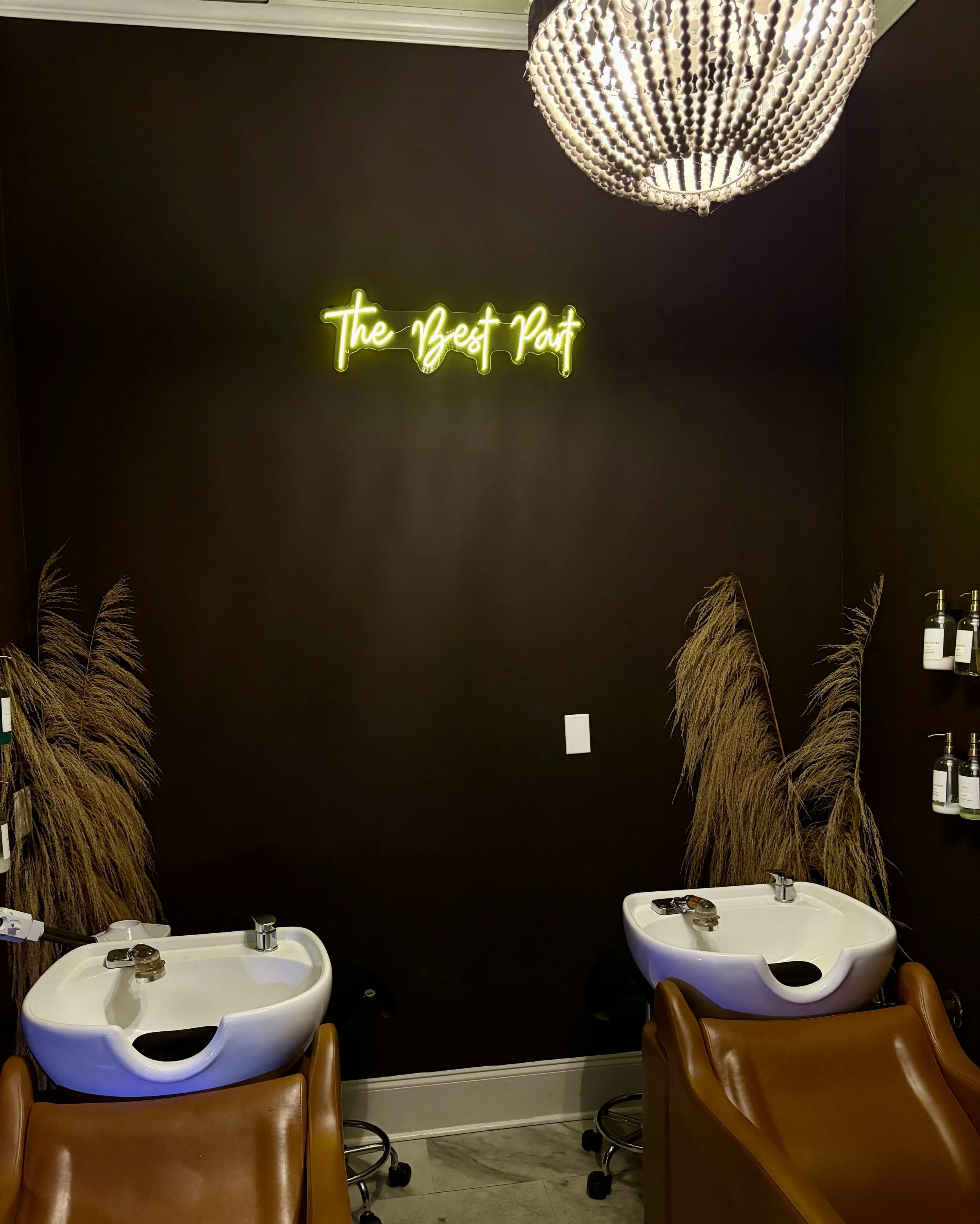 Salon wash stations with hair sinks and chairs, decorative pampas grass, a neon sign that reads 'The Best Part', a chandelier, and shelves with hair products on a dark painted wall.