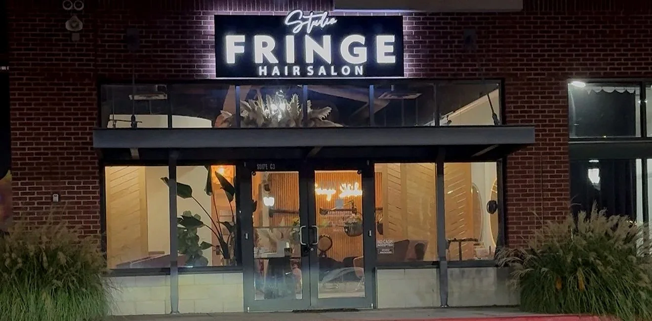 Exterior of a hair salon named 'Studio Fringe' with a brick facade, large glass windows, and a lit sign above the entrance. Inside, visible are chairs and plants.
