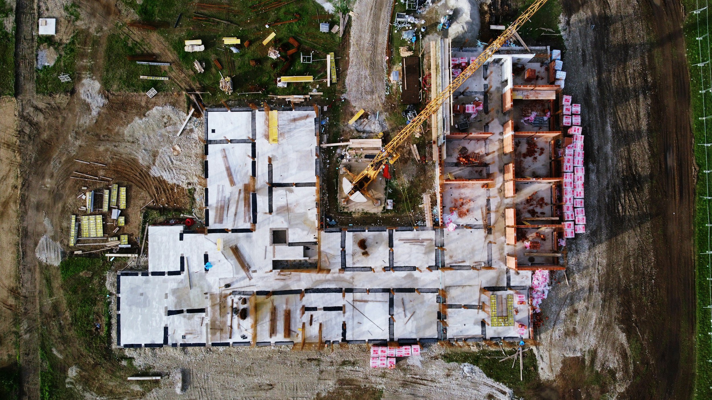 Vogelperspektive auf den Bau eines Gebäudes mit sichtbaren Fundamenten und einem Kran, umgeben von einem unbefestigten Baugrund und Grünflächen.