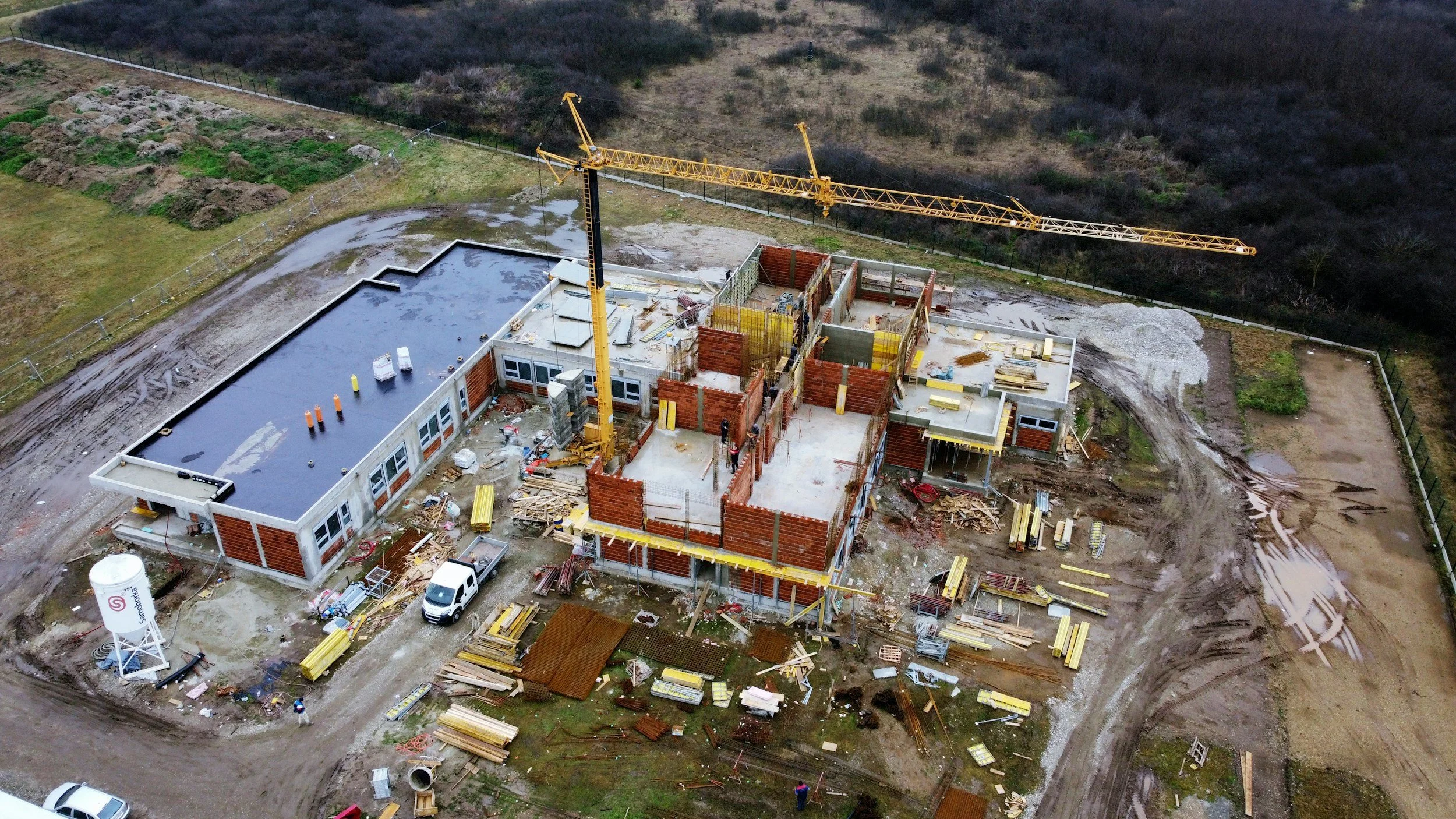 Eine Baustelle mit einem halbgebauten Gebäude, Kran und Baumaterialien, umgeben von unbefestigten Wegen und einem umgrenzten Gelände in einer ländlichen Umgebung.
