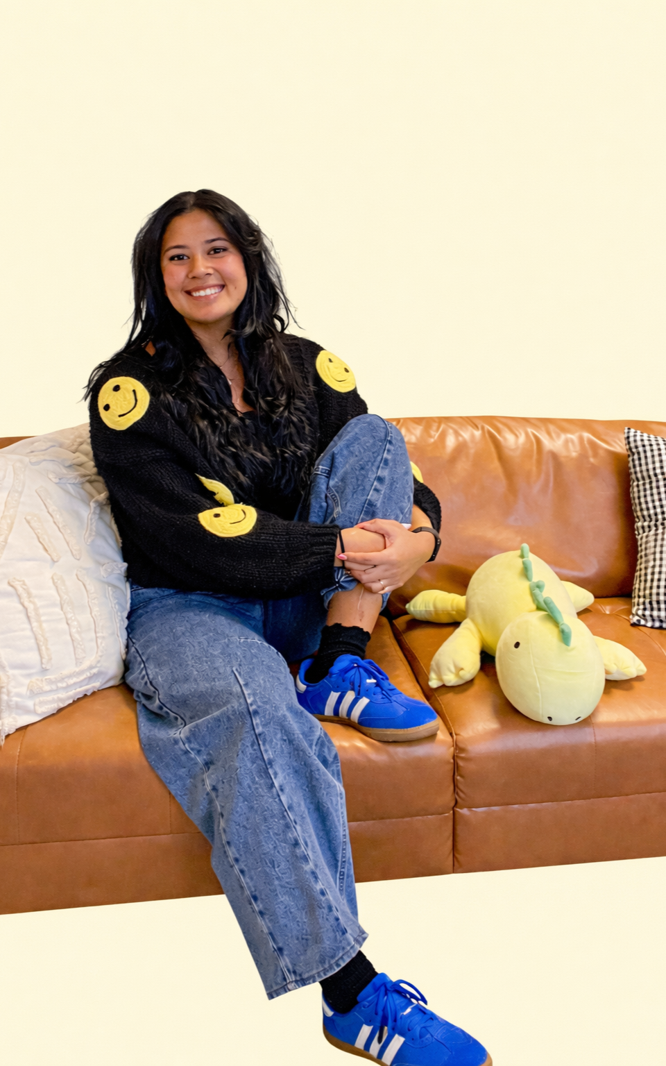 A young woman with long black hair sitting on a brown leather couch, smiling, wearing a black sweater with yellow smiley face patches, blue jeans, and blue Adidas sneakers, with a plush yellow dinosaur toy on the couch beside her.