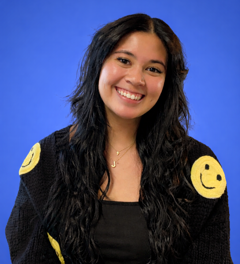 A young woman with long, wet black hair smiling against a blue background. She wears a black top and a black knitted sweater with yellow smiley face patches on the shoulders, along with layered necklaces.