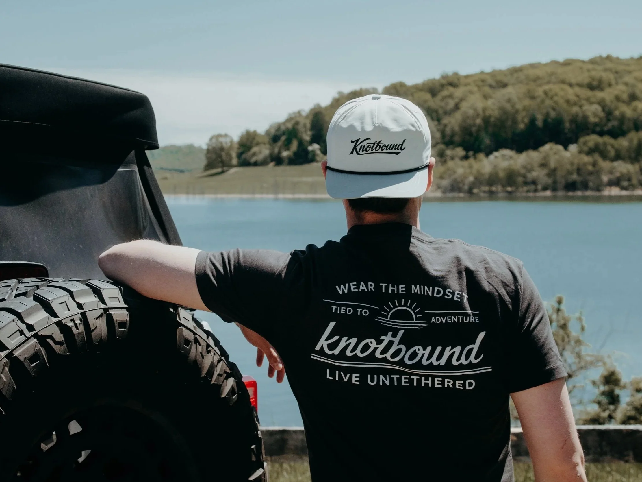 Back view of a person wearing a white cap with the logo 'Knotbound' and a black T-shirt with the text 'Wear the mindset tied to adventure knotbound live untethered' while leaning on a vehicle. A large tire is visible in the foreground with a lake and green hills in the background.