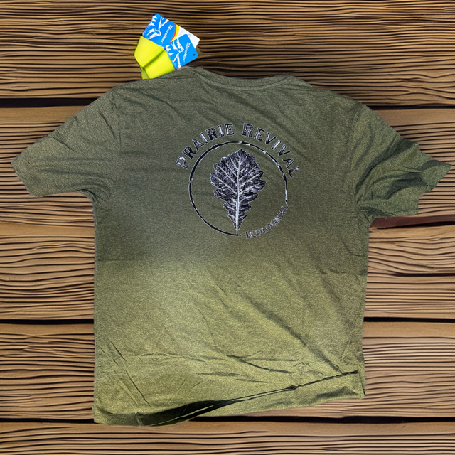 Flatlay photo of a green heathered performance t-shirt on a dark wood background, showcasing the full Prairie Revival custom back print.