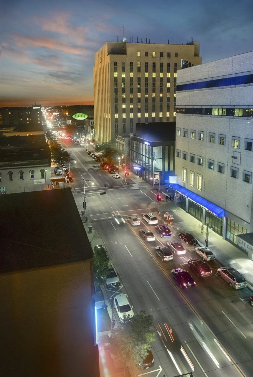 City street during sunset with tall buildings, traffic, and parked cars, with some building lights illuminating.