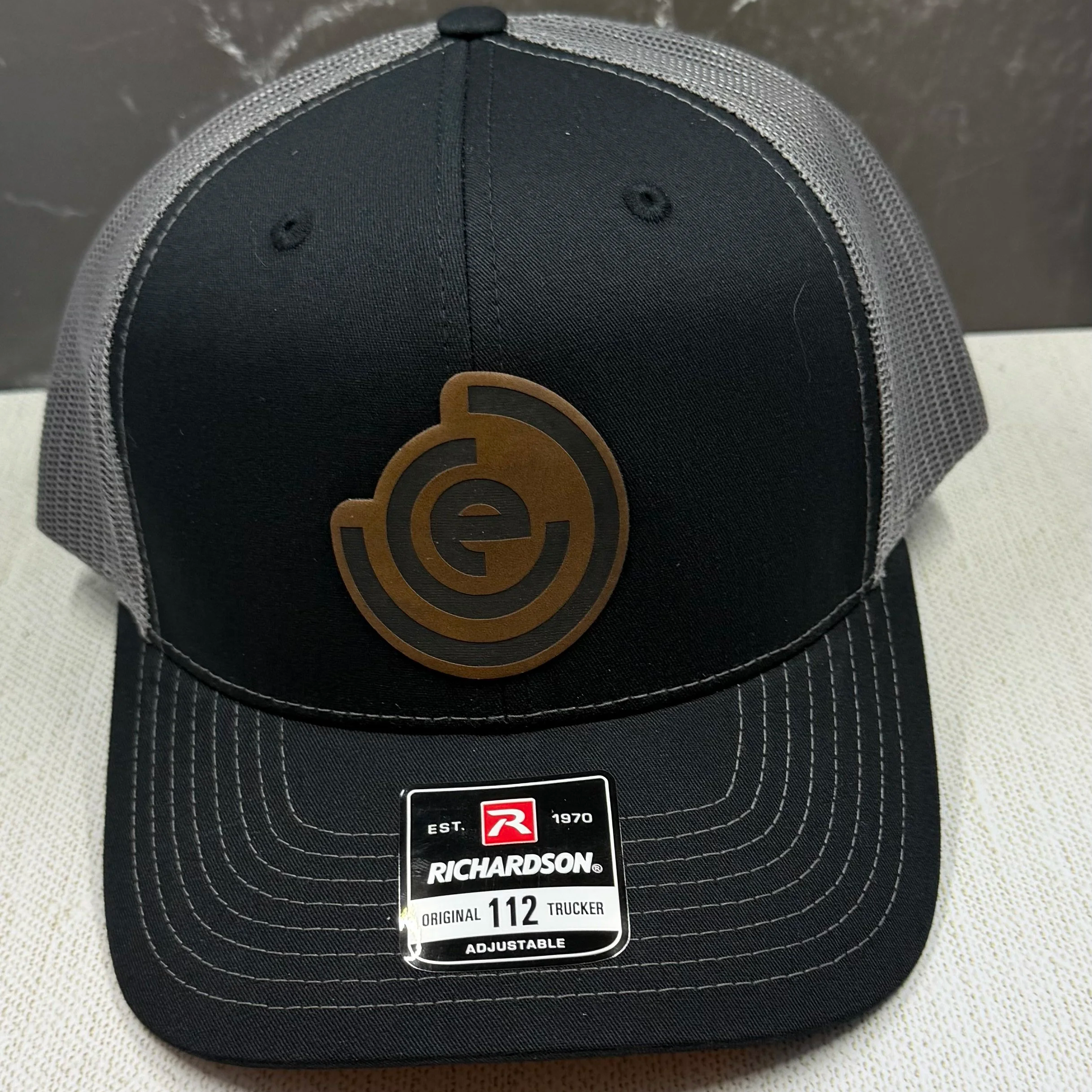 Black Richardson trucker cap with gray mesh sides, a leather patch on the front, and a White tag indicating size 112 adjustable fit.