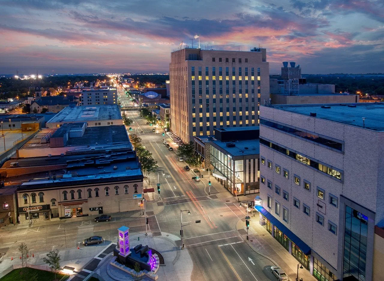 City street at dusk with tall buildings, streetlights, and cars. The sky has pink and purple clouds. There are visible light trails from moving vehicles and a purple-lit monument in the foreground.