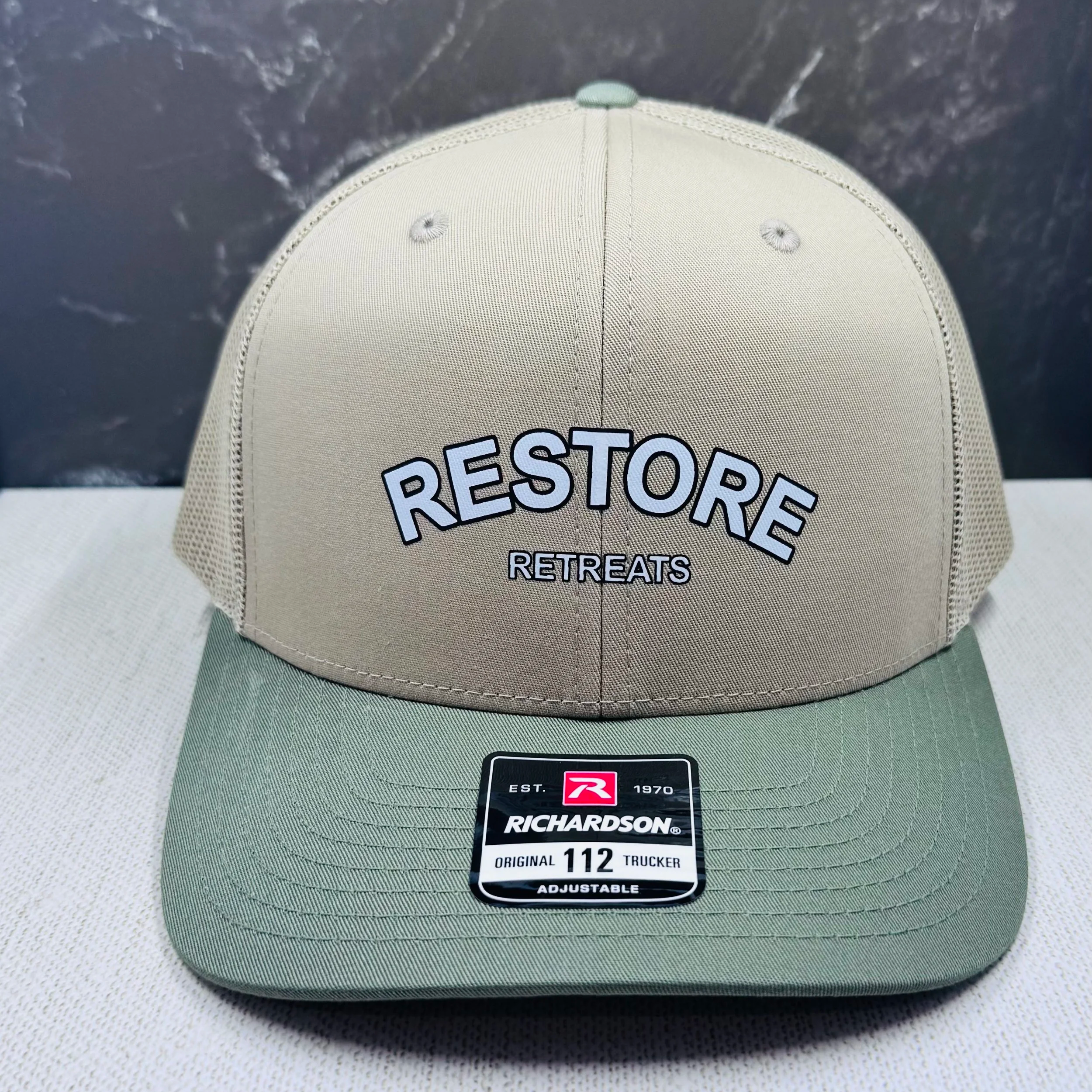 Beige trucker hat with embroidered text and a sticker on the brim.