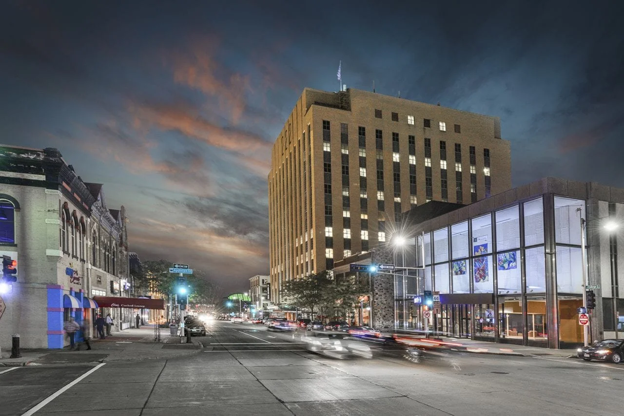 City street scene during dusk with a tall, illuminated office building and storefronts on either side, traffic and pedestrians in motion, and a partly cloudy sky with colorful clouds.