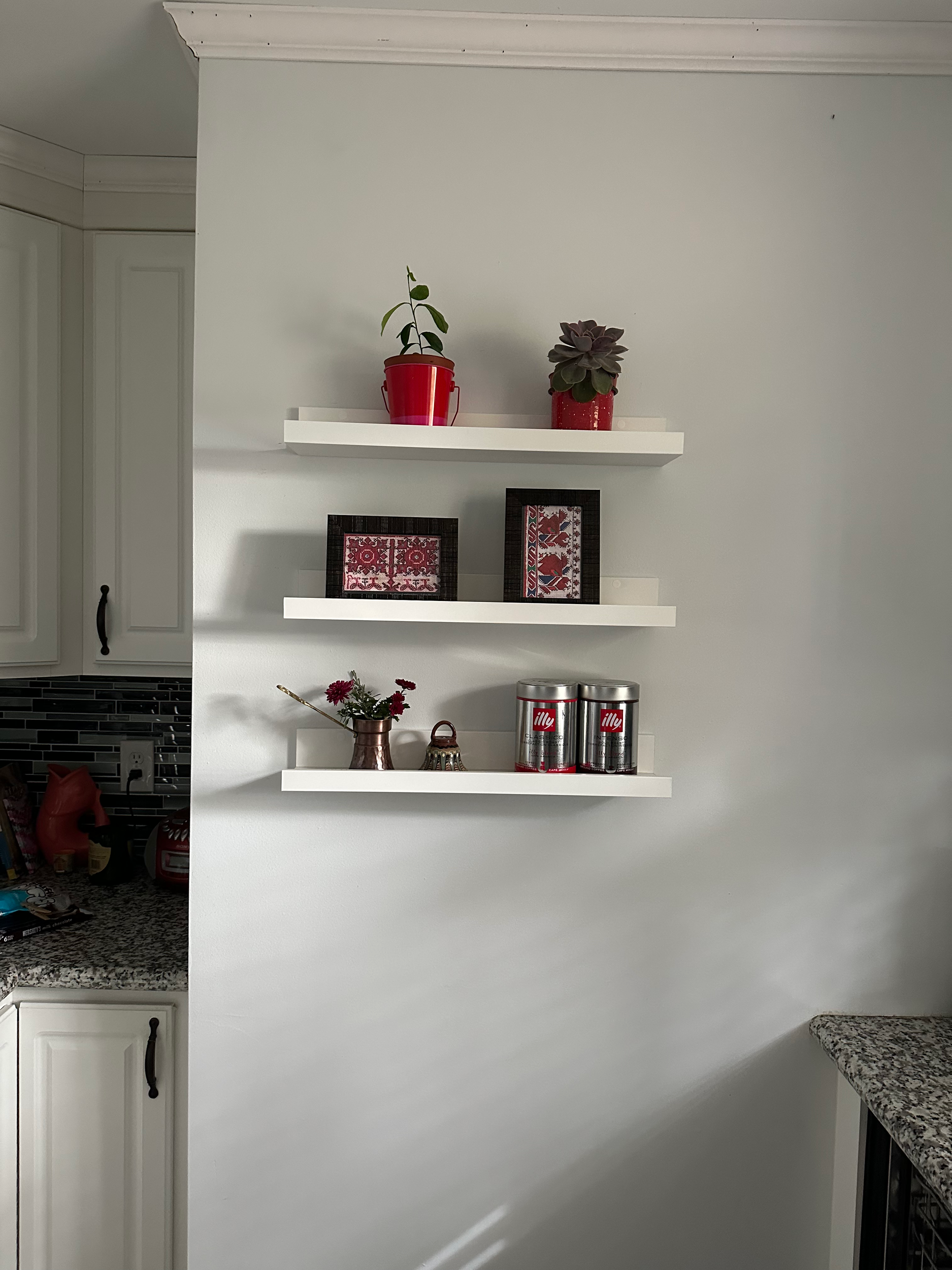 Three white floating shelves on a light gray wall with plants, framed artwork, flowers in a vase, and canned coffee containers. Part of a kitchen with granite countertops and cabinets visible on the left.