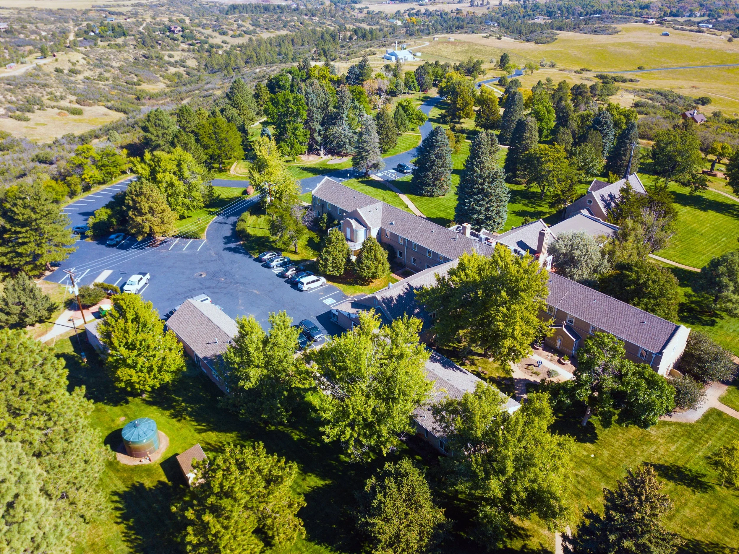 Aerial view of sacred heart retreat house, parking lots, and green trees in a semi-rural setting. Fields and rolling hills are visible in the background.