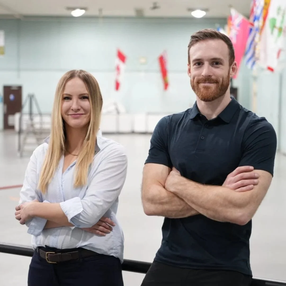 A man and woman standing with arms crossed, smiling, in a large indoor space with international flags in the background.
